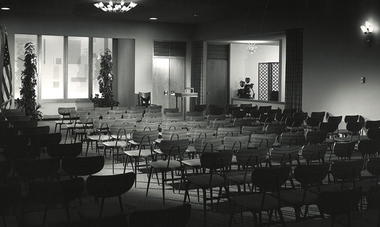 Black and white photo of an empty auditorium with rows of chairs facing a podium and microphone. Large windows, potted plants, and a US flag are visible in the background.