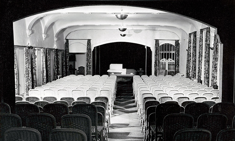 Black-and-white photo of an empty theater or auditorium with rows of chairs facing a stage. The stage has a table and a sofa, and is framed by curtains and arched architecture.