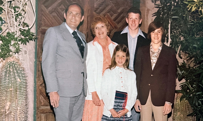 A family of five poses together, dressed in vintage clothing. Two adults stand with three children—two boys and one girl—in front of a wooden door and surrounded by plants. The young girl holds a book and smiles.