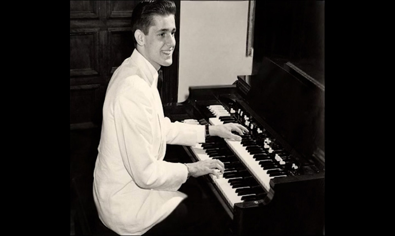 A young man in a white jacket sits at an organ, smiling as he plays. The photo is black and white, with wooden paneling and part of a blank wall in the background.