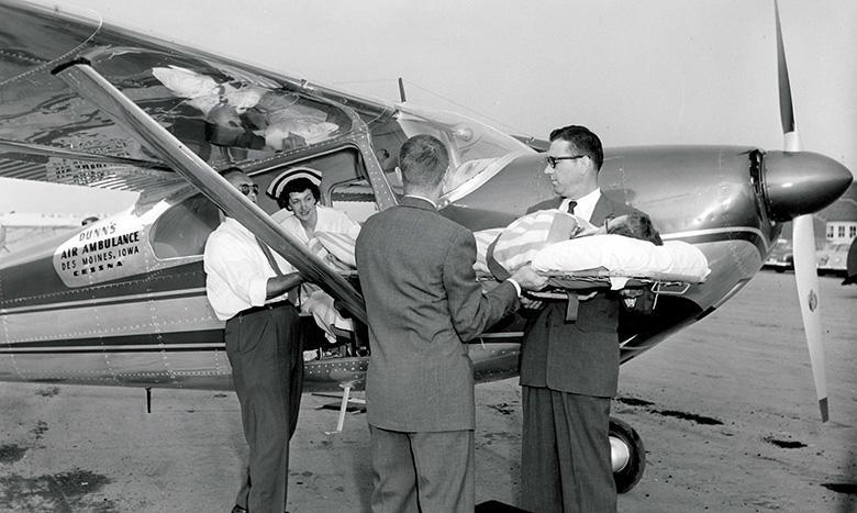 Two men and a nurse load a patient on a stretcher into a small airplane labeled “Davis Air Ambulance, Des Moines, Iowa.” The scene appears to take place at an airport in the mid-20th century.