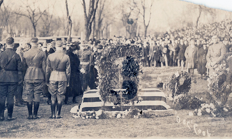 A black-and-white photo shows a funeral scene with soldiers in uniform standing near a flag-draped casket. Large wreaths and flowers surround the casket, and a crowd of mourners stands in the background.