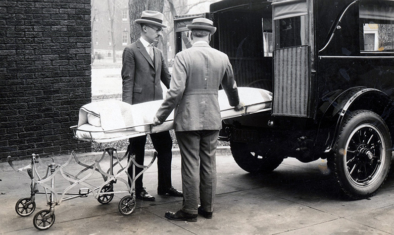 Two men in suits and hats load a covered stretcher into the back of a vintage hearse. An empty, folded stretcher is on the ground nearby, and the scene appears to be from the early 20th century.