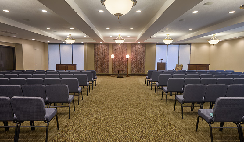 Rows of empty blue chairs face a stage with two podiums and decorative lamps in the well-lit, beige-carpeted Iles Grandview Park Chapel conference or meeting room.