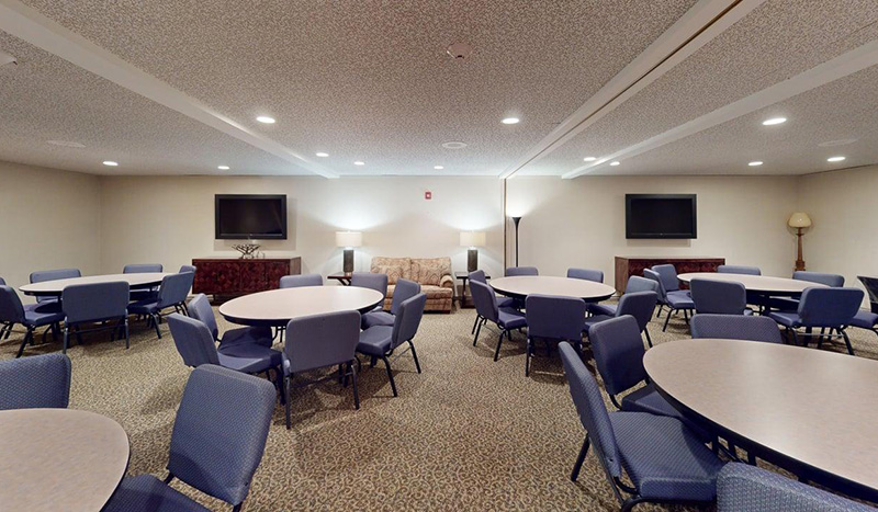 A conference room at Iles Grandview Park Chapel features several round tables surrounded by blue chairs, two large wall-mounted TVs, a beige sofa, lamps, and light-colored walls with patterned carpet flooring.