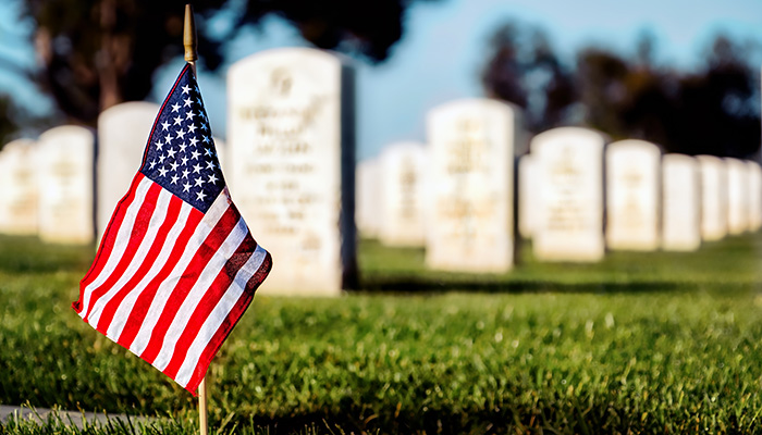 An American flag on a small pole stands before rows of white headstones in a cemetery, symbolizing honor and remembrance for those who served and may be eligible for Veterans Funeral Benefits.