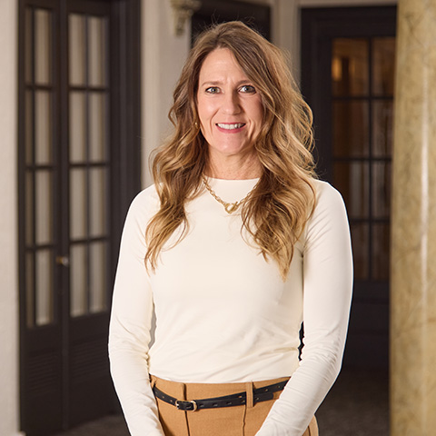 A woman with long wavy hair, wearing a white long-sleeve top and tan pants with a black belt, stands indoors smiling warmly, reminiscent of caring staff, with dark-paneled doors and a marble column in the background.