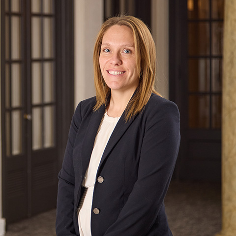 A woman with straight, light brown hair wearing a dark blazer and white blouse stands indoors, smiling warmly at the camera—reflecting the caring staff often found in professional settings—posed in front of glass-paneled doors and beige walls.