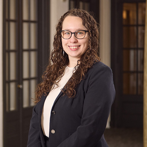 A woman with long curly brown hair and glasses, wearing a black blazer and white top, stands smiling in a hallway with glass-paneled doors and warm lighting, reflecting the welcoming atmosphere created by the caring staff.