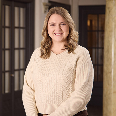 A young woman with blonde wavy hair is smiling, wearing a cream-colored cable-knit sweater, standing indoors in front of dark wooden doors and a marble column, embodying the warmth you’d expect from caring staff.