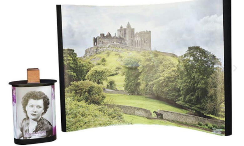 A photo display features a black-and-white portrait of a woman on a small cylindrical holder, evoking life celebrations, next to a curved backdrop showing a hilltop castle surrounded by greenery and stone walls under a cloudy sky.