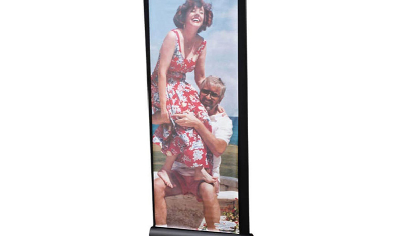 A freestanding banner displays a photo of a smiling woman in a red floral dress sitting on a man's shoulders outdoors, both appearing happy and playful under a bright sky—capturing the spirit of life celebrations.