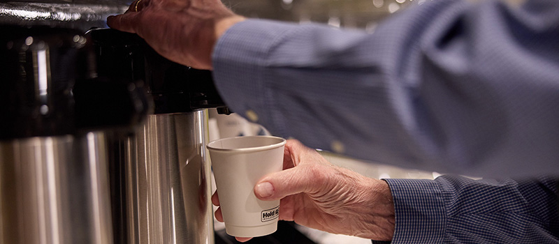 A person in a blue shirt dispenses coffee from a stainless steel coffee urn into a white paper cup at Sundberg Kirkpatrick Chapel, holding the cup under the spout with one hand and pressing the lever with the other.