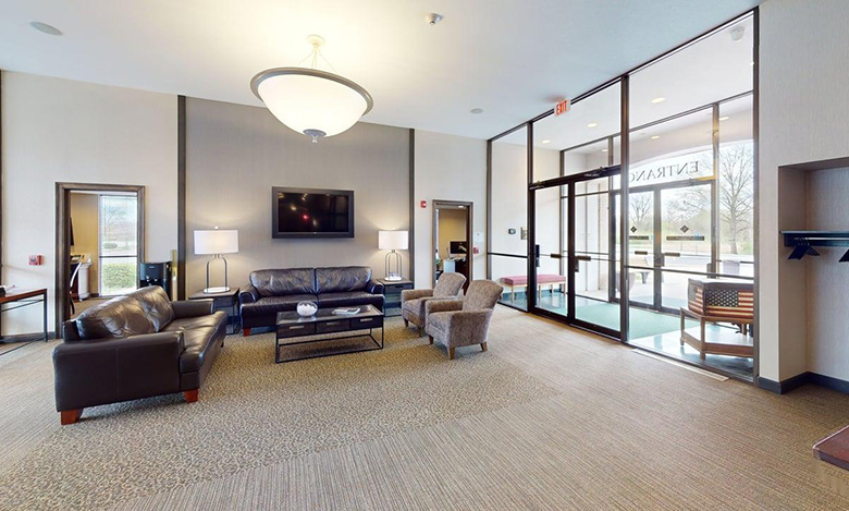 A modern lobby at Iles Grandview Park Chapel features leather sofas, armchairs, a coffee table, lamps, and a wall-mounted TV. Large glass doors and windows provide natural light and an outdoor view in a welcoming, neutral-toned setting.