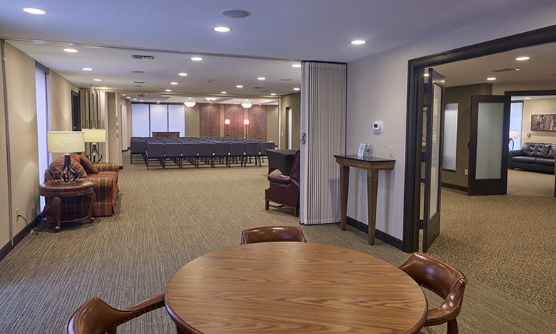 A spacious, well-lit room at Iles Grandview Park Chapel features a round wooden table and chairs in the foreground, armchairs and a lamp on the left, and rows of chairs facing a wall. Separate rooms are visible on the right.