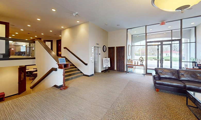 A spacious lobby at Iles Grandview Park Chapel features carpeted floors, a dark brown leather couch, glass doors and windows, a staircase with a wooden banister, and a sign near the entrance. Bright lighting and neutral walls create a welcoming atmosphere.