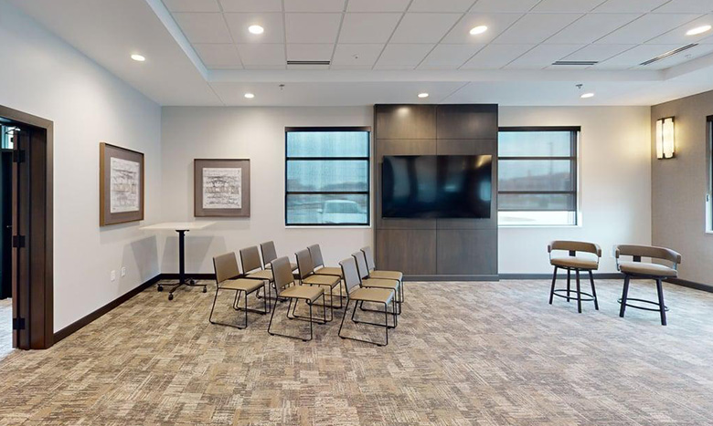 Modern conference room at Jordan Creek Chapel with rows of chairs facing a large wall-mounted TV, two armchairs to the side, framed art on the walls, and carpeted flooring. The room is well-lit with windows and ceiling lights.