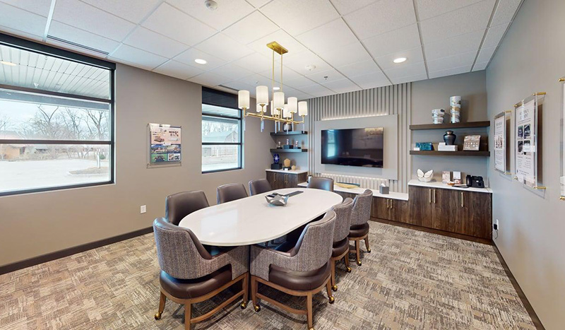Modern conference room with an oval white table, eight gray chairs, a wall-mounted TV, shelves with decor, and large windows at Jordan Creek Chapel. Framed posters adorn the walls as natural light fills the space, highlighting the beige carpeting.