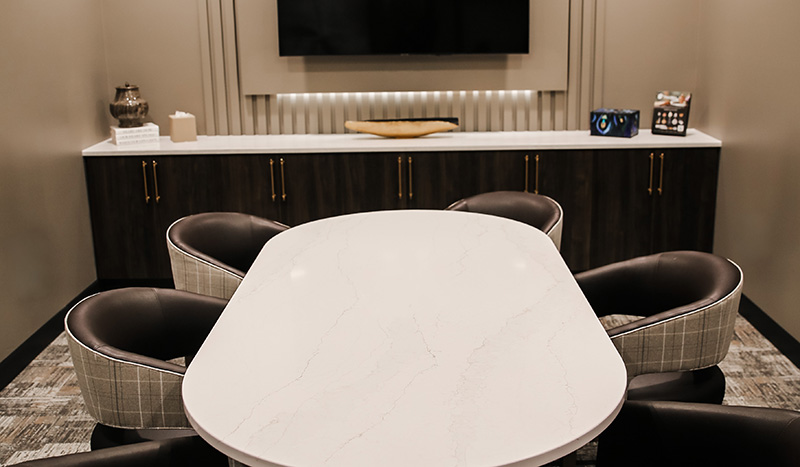 A modern meeting room at Jordan Creek Chapel features a white marble oval table, five curved plaid chairs, and a dark cabinet along the back wall beneath a large TV and decorative items.