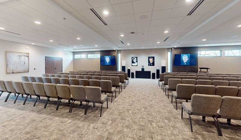 A modern, empty chapel with rows of gray chairs facing a podium. Three large screens at the front display a portrait. Jordan Creek Chapel features neutral walls, ceiling lights, and a large abstract painting on the left wall.