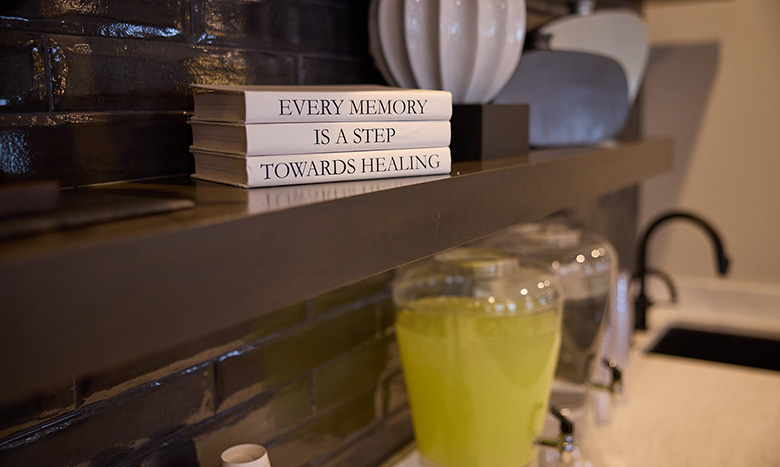 A close-up of a modern kitchen shelf displays three stacked books with spines reading “Every memory is a step towards healing.” Nearby, a glass dispenser filled with a yellow drink brightens the countertop—like sunlight streaming into Jordan Creek Chapel.