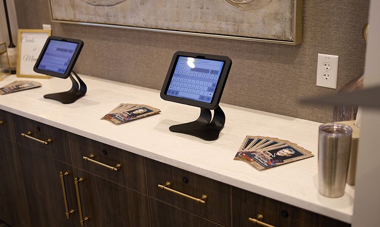 Two touchscreen tablets on stands sit on a counter with pamphlets and a metal tumbler nearby at Jordan Creek Chapel. The counter has gold handles, and a nearby outlet is visible on the wall.