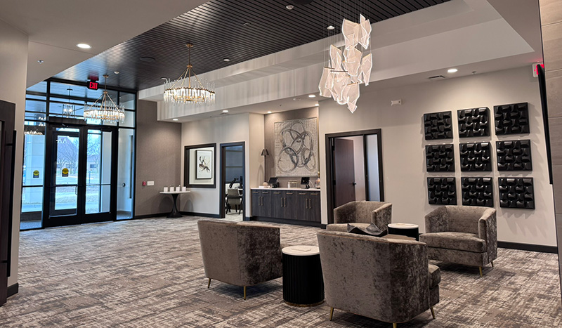 Modern lobby at Jordan Creek Chapel with grey armchairs, round tables, wall art, and geometric chandeliers. Large windows fill the space with natural light. Grey carpet and neutral walls create an elegant, inviting atmosphere.