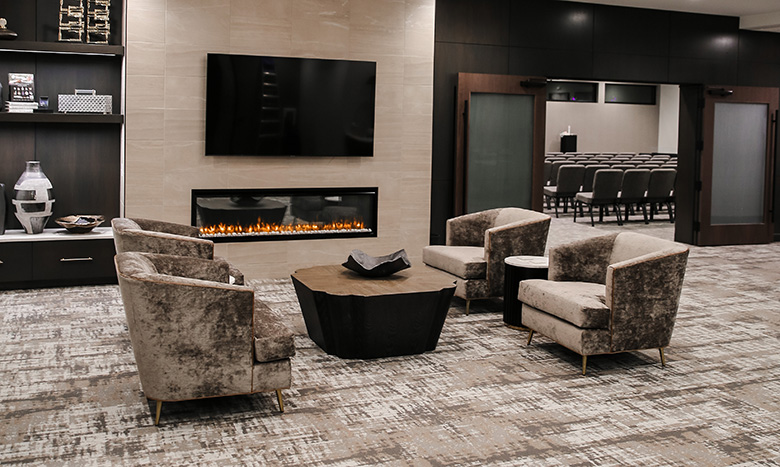 Modern lounge area with four plush armchairs around a wooden coffee table, set before a wall-mounted TV and long fireplace. Shelving and decor are visible, with a view into the Jordan Creek Chapel conference room lined with rows of chairs.