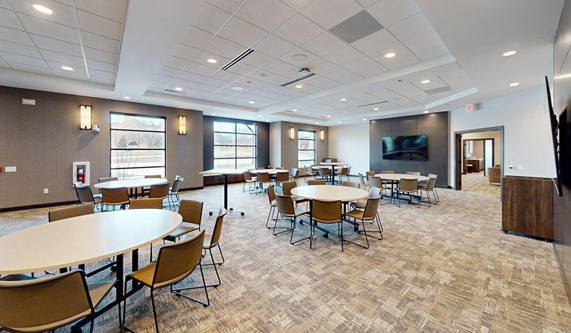 A spacious, modern meeting room at Jordan Creek Chapel with round tables, chairs, a carpeted floor, large windows, wall-mounted TV, and neutral-toned walls, all illuminated by ceiling lights.