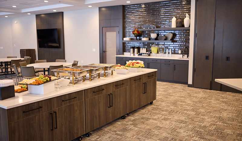 A modern conference room at Jordan Creek Chapel features a buffet table with trays of food, fruit, and drinks. Behind the counter, shelves display decorative vases and cups, while tables with chairs are arranged for dining.