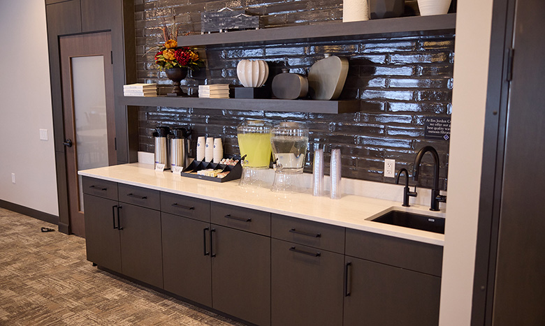 A modern refreshment station at Jordan Creek Chapel features dark cabinets, open shelves with vases and books, coffee dispensers, lemonade, cups, and a sink, all set against a glossy dark tile backsplash.