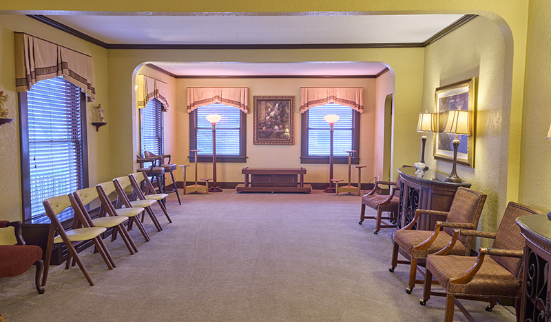 A spacious, warmly lit room with yellow walls, rows of chairs along each side, windows with blinds and valances, lamps, framed art, and a sideboard with decor—Sundberg Kirkpatrick Chapel creates a formal yet inviting atmosphere.