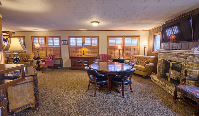 A cozy living room with carpeted floor, a round table with four chairs, sofas, armchairs, lamps, and a stone fireplace evokes the inviting atmosphere of Sundberg Kirkpatrick Chapel. Multiple windows with wooden shutters fill the space with natural light.